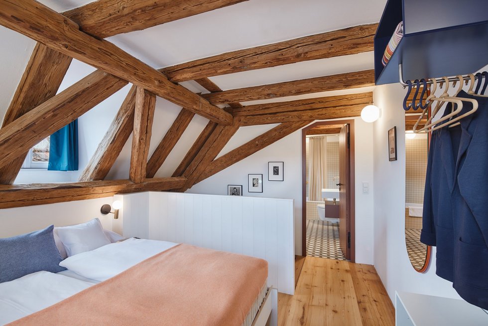 Heller Raum mit sichtbaren Deckenbalken, großem Doppelbett und Blick in das moderne Badezimmer